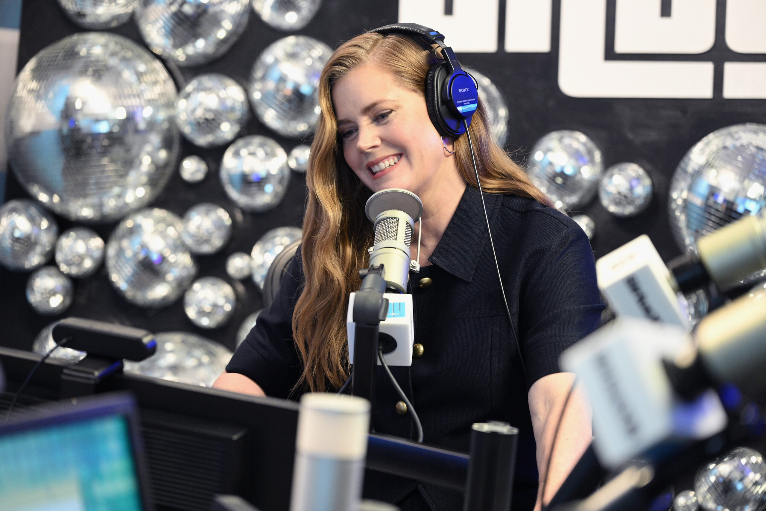 Amy Adams, a woman with long, wavy hair wearing headphones smiles while speaking into a microphone in a radio studio decorated with shiny disco balls. Listen to Radio Andy on SiriusXM.