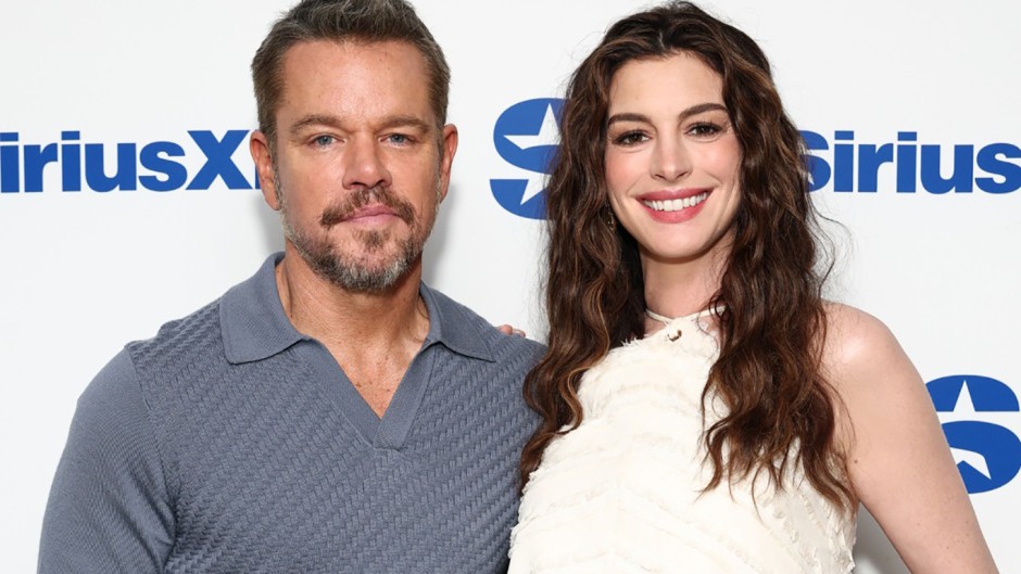 Matt Damon and Anne Hathaway stand together smiling in front of a SiriusXM logo wall. Matt wears a gray polo shirt and has a beard, while Anne wears a sleeveless white dress with wavy brown hair. This is to promote an interview about The Odyssey