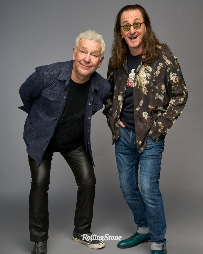 Alex Lifeson and Geddy Lee, two smiling men pose in casual clothes against a gray backdrop. Alex LIfeson leans forward with hands behind his back; Geddy Lee stands with hands in his pockets. The Rolling Stone logo appears at the bottom.